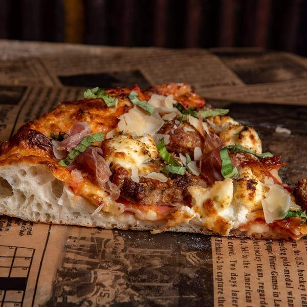 A close-up of a Ginny's pizza slice with meatballs, prosciutto, ricotta, and basil on newspaper.