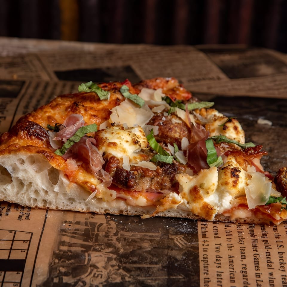 A close-up of a Ginny's pizza slice with meatballs, prosciutto, ricotta, and basil on newspaper.