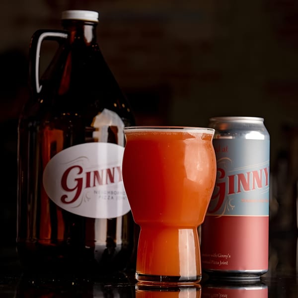 A Ginny's growler, a glass of orange beer, and a Ginny's beer can on a dark surface.