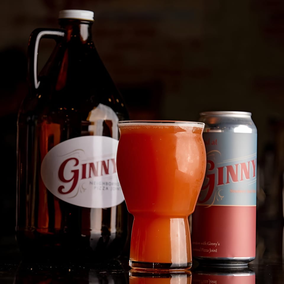 A Ginny's growler, a glass of orange beer, and a Ginny's beer can on a dark surface.