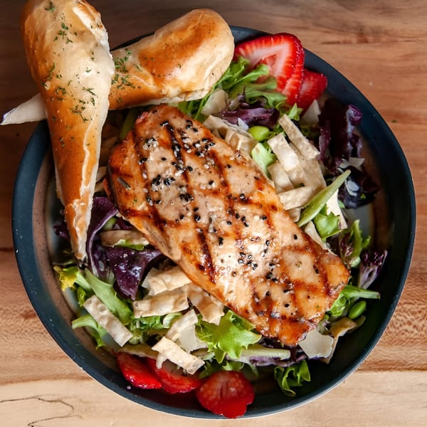 Grilled salmon salad with mixed greens, strawberries, and breadsticks in a dark bowl.