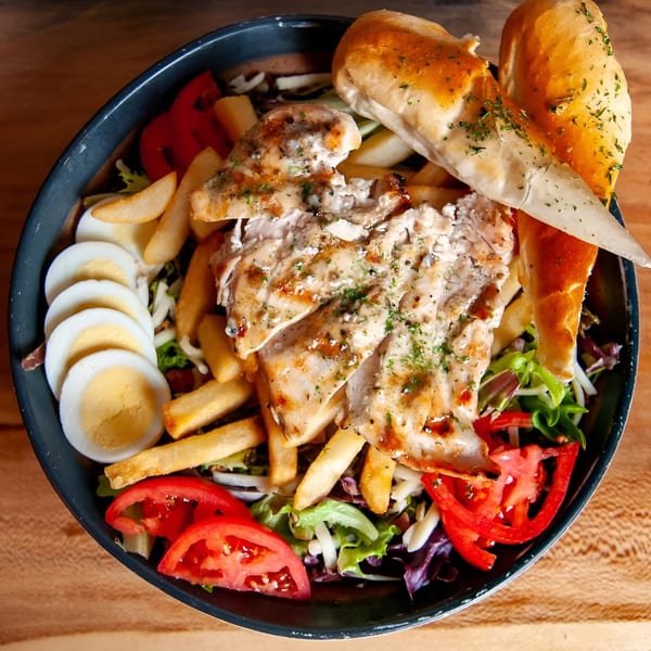 A large salad bowl with grilled chicken, french fries, sliced tomatoes, hard-boiled eggs, and bread.