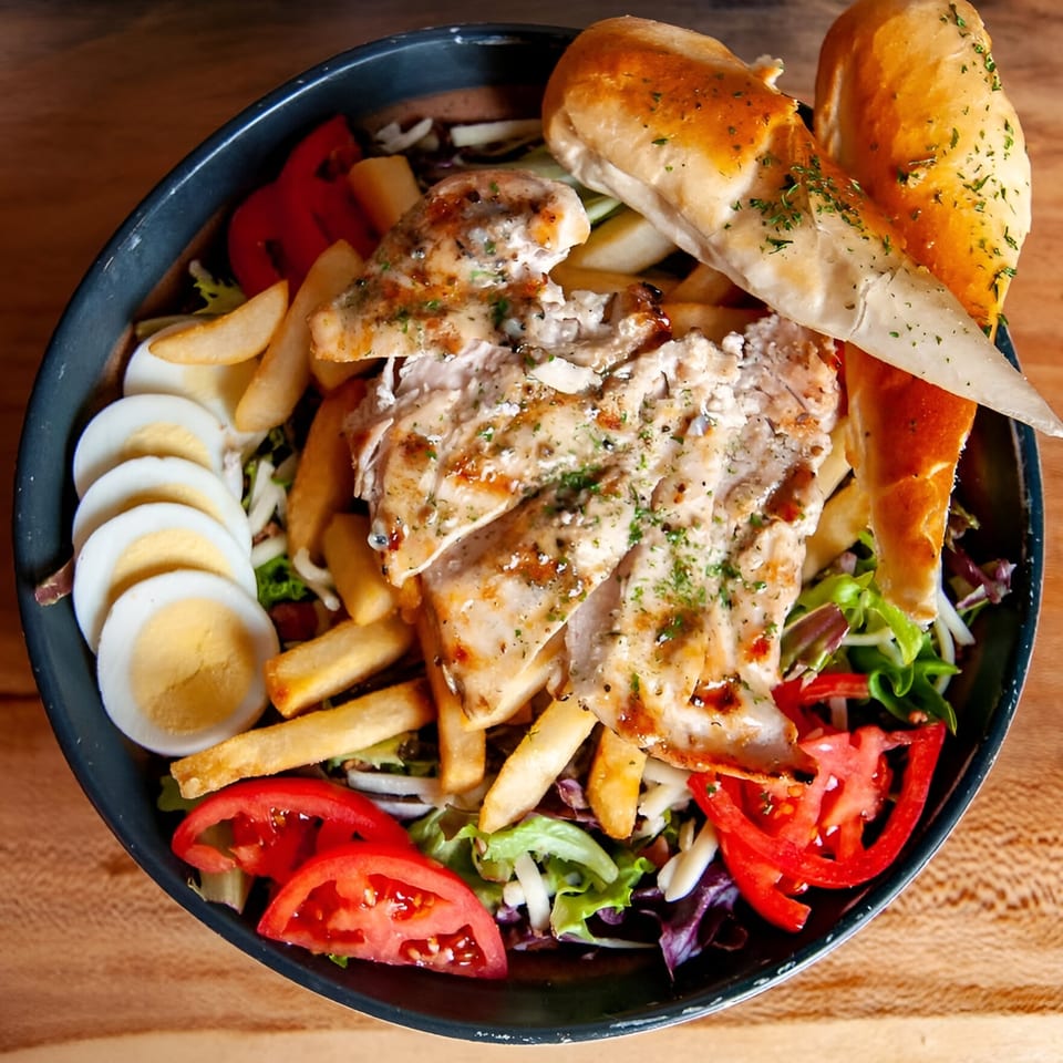 A large salad bowl with grilled chicken, french fries, sliced tomatoes, hard-boiled eggs, and bread.