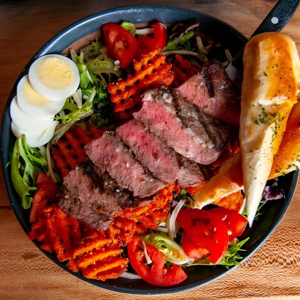 Steak salad with waffle fries, tomatoes, hard-boiled eggs, and bread in a black bowl.
